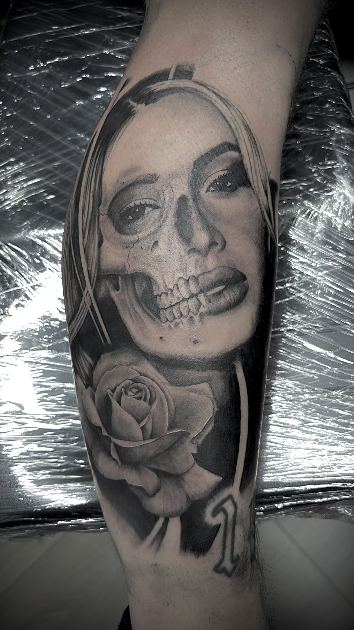 Realistische tattoo in zwart en grijs: een vrouwenportret dat half schedel is, met een roos eronder
